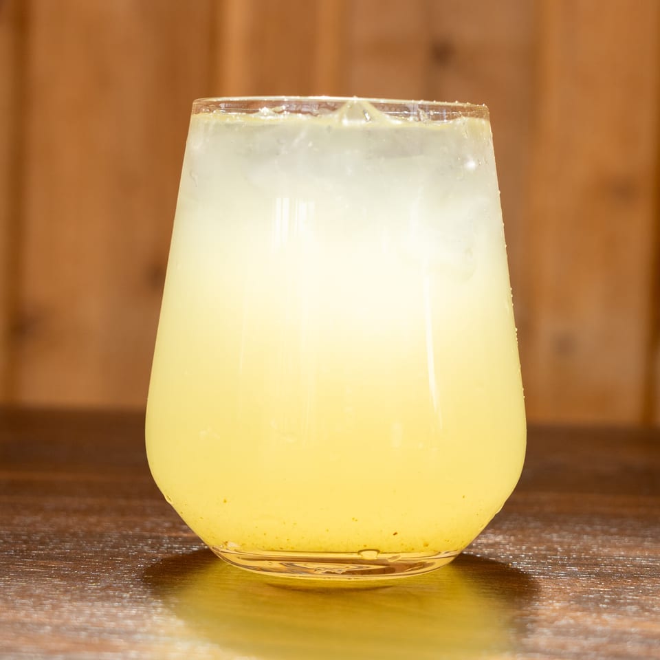 A chilled pale yellow beverage with ice fills a short glass on a wooden table against a warm brown background.