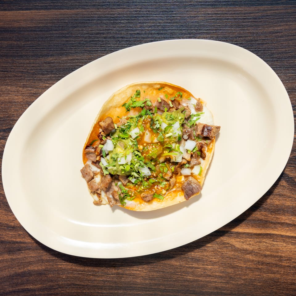 Tostada topped with grilled meat, white onion, cilantro, green guacamole, and orange salsa on a white plate.