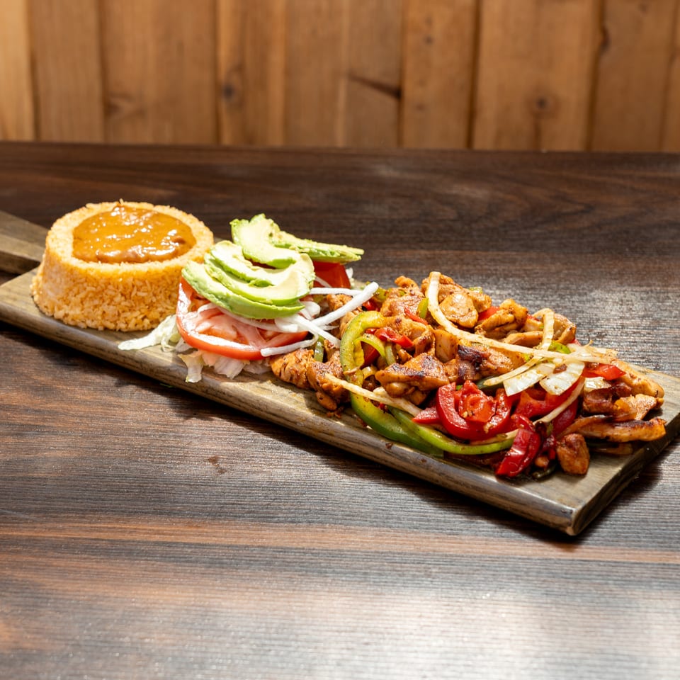 Grilled chicken with peppers, onions, avocado, tomato salad, and rice served on a wooden platter.