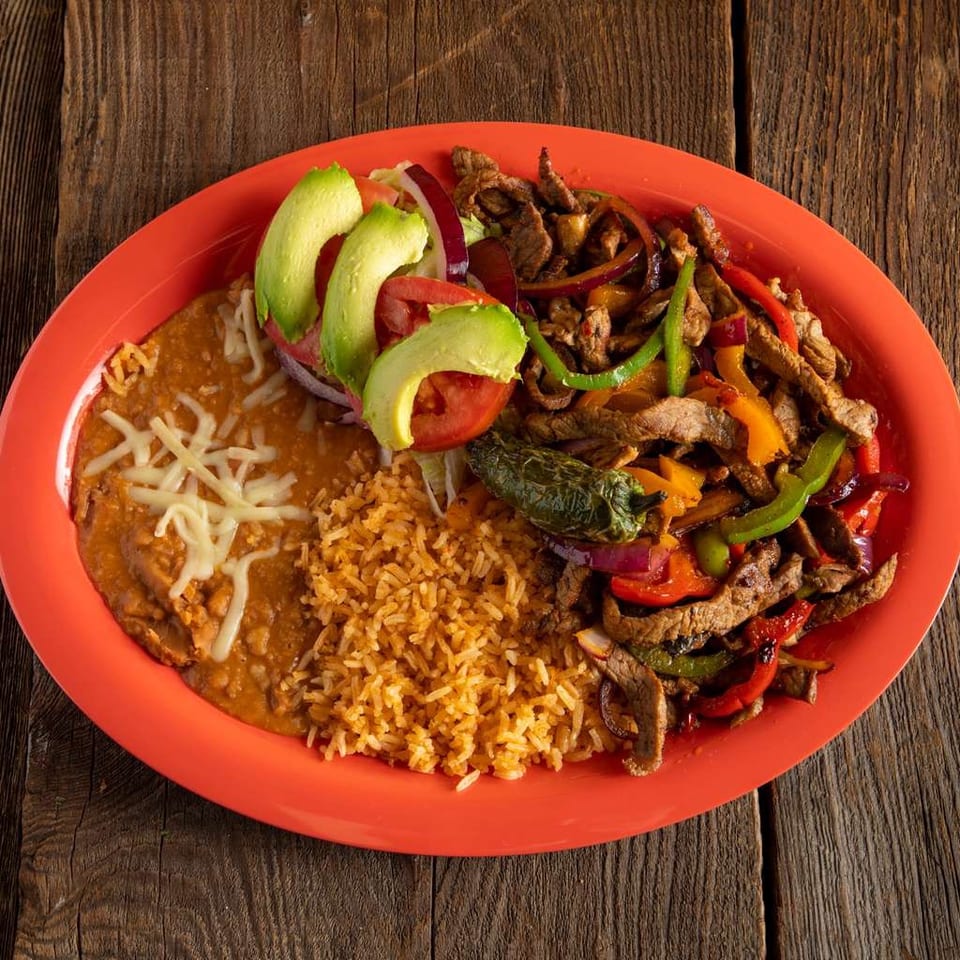 Mexican platter with steak fajitas, peppers, onions, rice, refried beans, melted cheese, avocado, and tomatoes on a red plate.