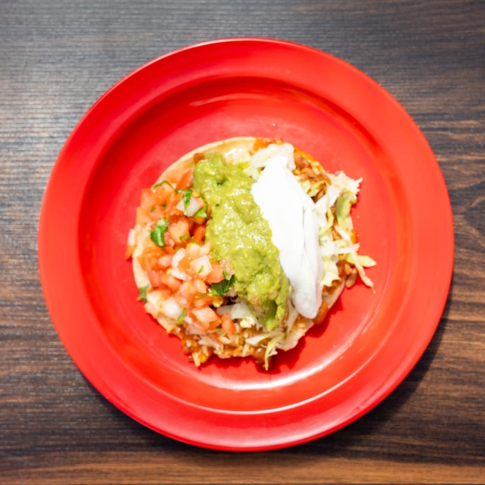 Mexican tostada topped with guacamole, sour cream, shredded lettuce, beans, and fresh pico de gallo on a red plate.