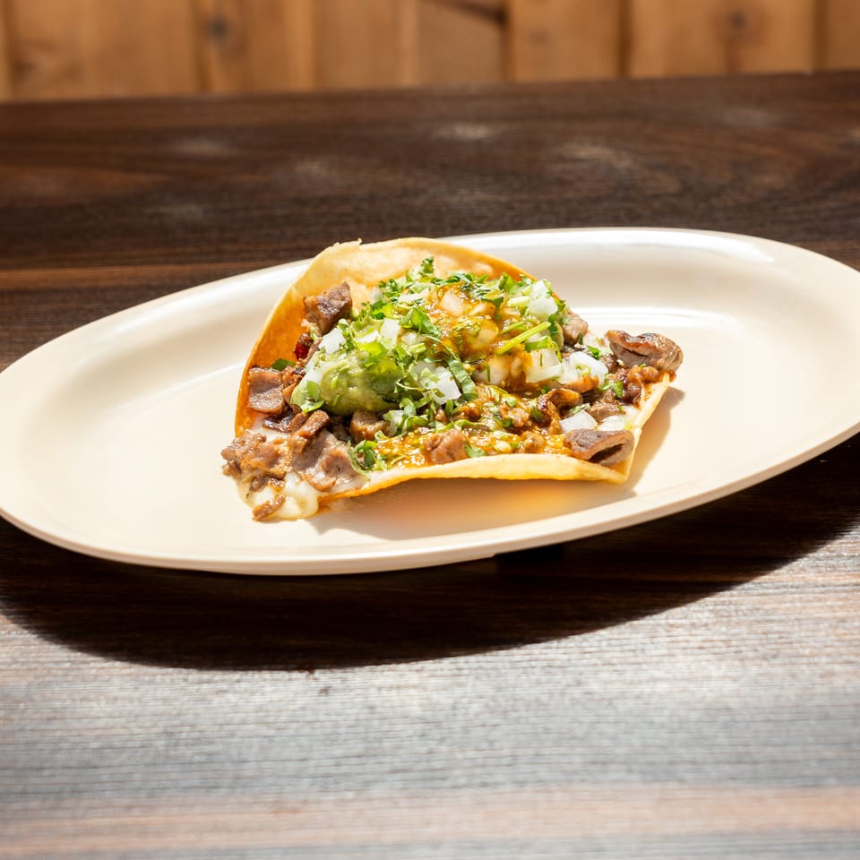 Loaded taco with grilled meat, onions, cilantro, guacamole, and salsa on a white plate atop a wooden table.
