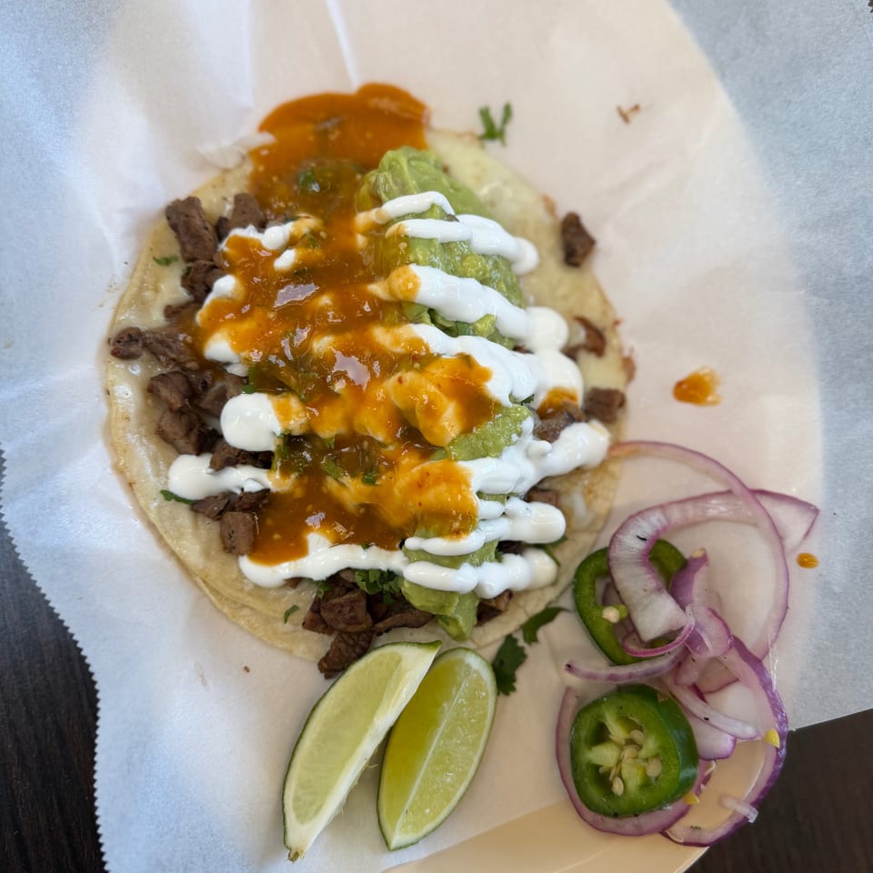 Steak tacos topped with guacamole, crema, and orange salsa, served with lime wedges, sliced jalapeños, and red onions.