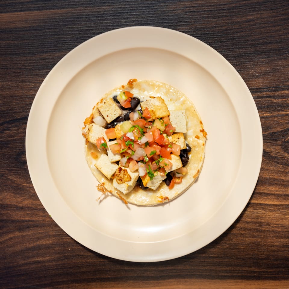 Plate of nachos topped with black beans, diced tomatoes, onions, cilantro, and golden tofu pieces on a wooden table.