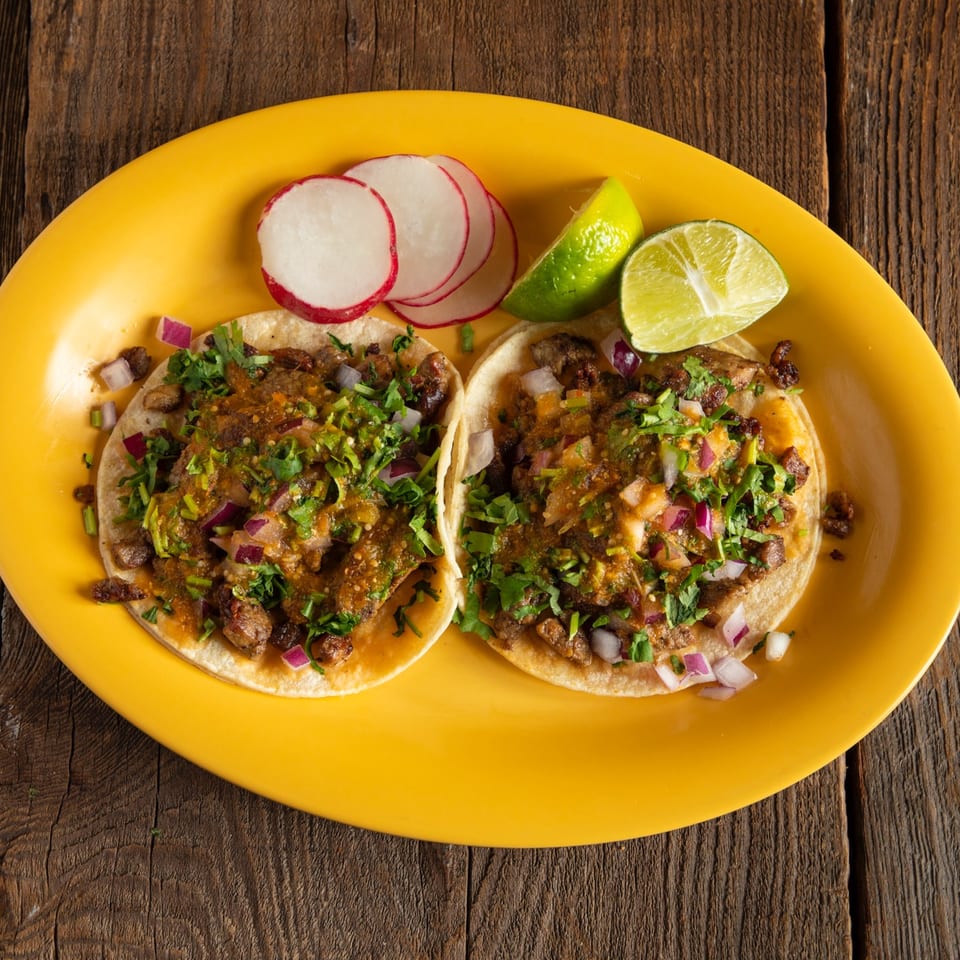 Two tacos topped with seasoned meat, salsa, cilantro, and onions on a yellow plate with radishes and lime wedges.