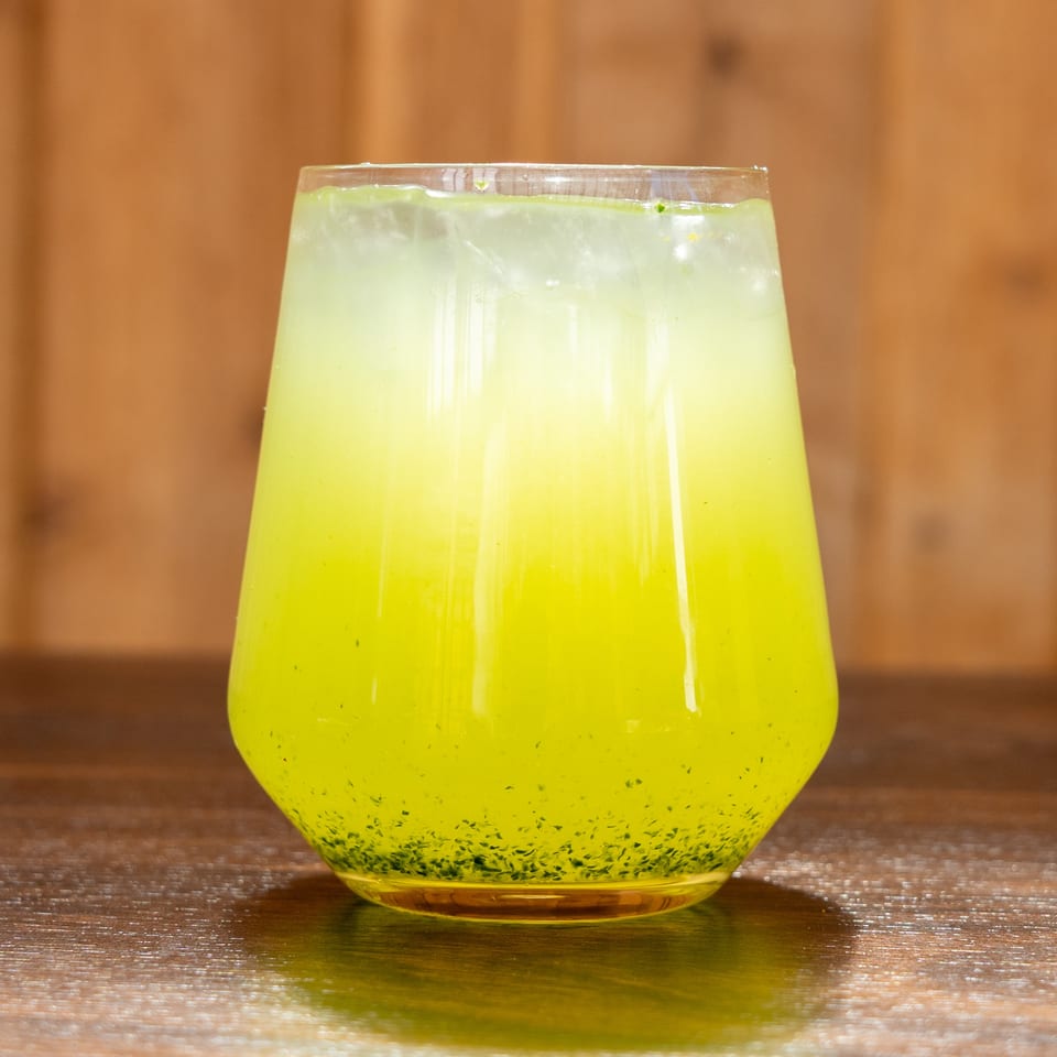 Bright green iced drink in a clear glass, garnished with herbs, sits on a wooden table against a warm brown background.