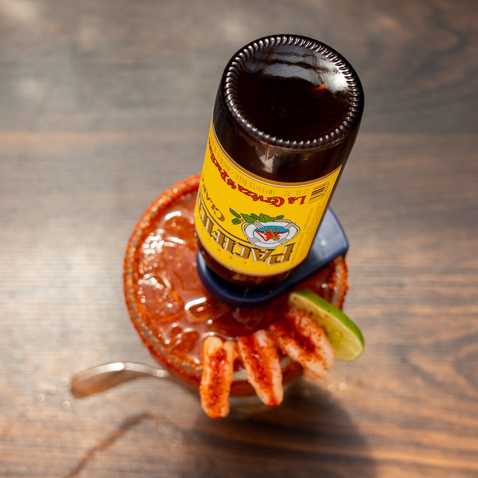 Bottle of La Guera hot sauce atop a chili-coated dish with shrimp, lime wedge, and spoon on a wooden table.