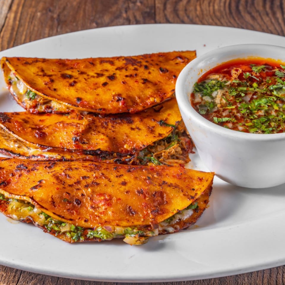 Three crispy orange tacos filled with meat and herbs sit beside a bowl of red salsa on a white plate.