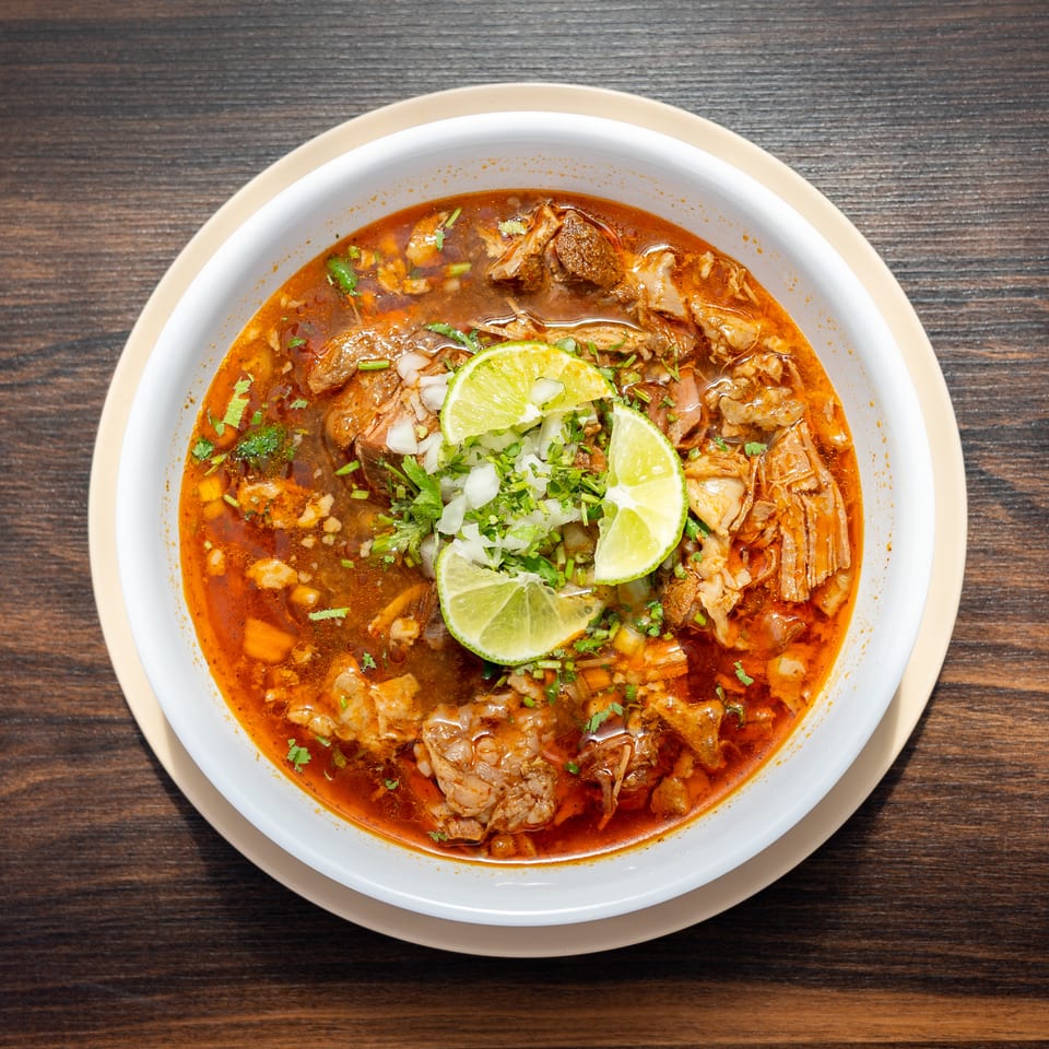 Bowl of spicy red broth with tender meat, chopped onions, cilantro, and lime wedges on a wooden table.