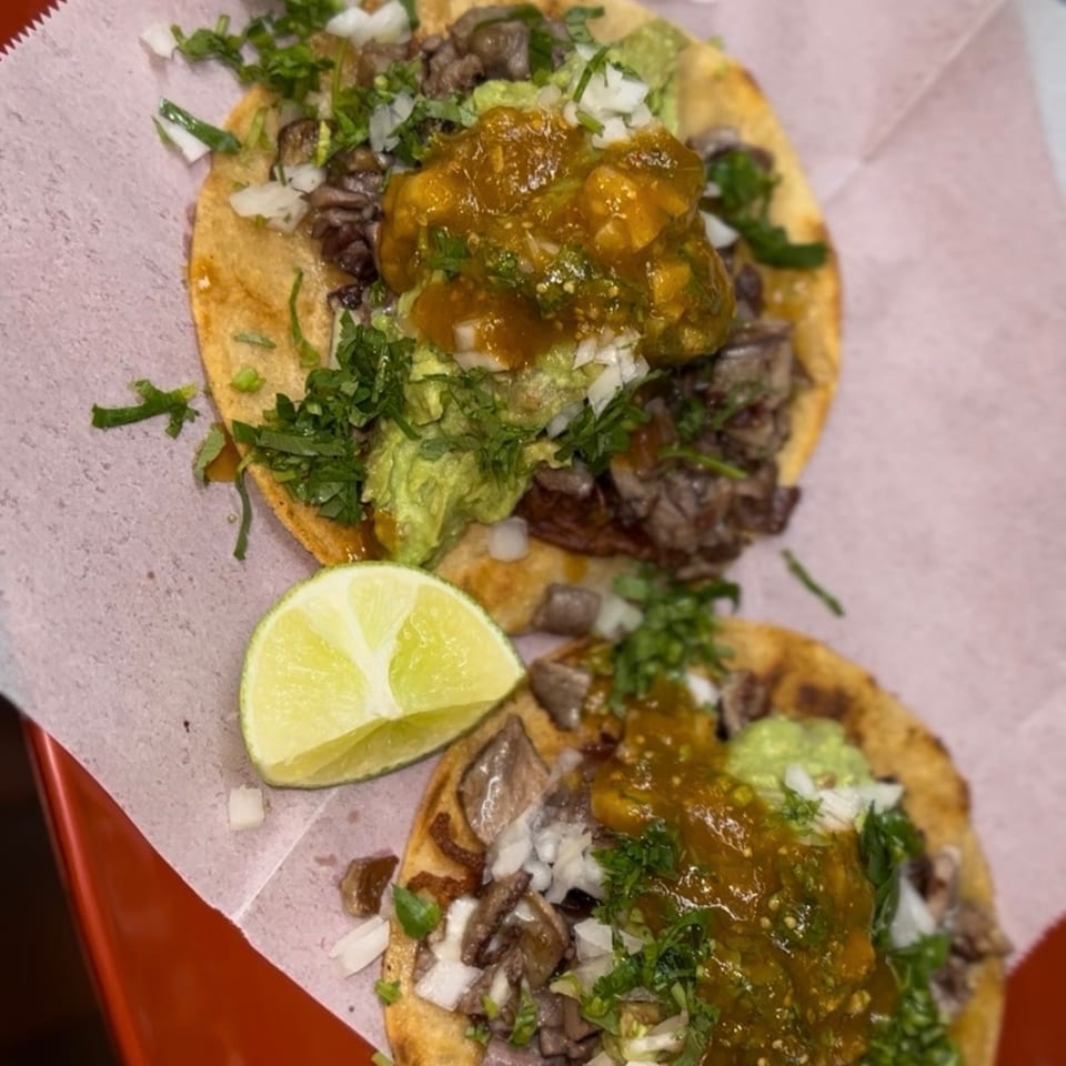 Two tostadas topped with grilled meat, guacamole, salsa, diced onions, and cilantro, served with a lime wedge.